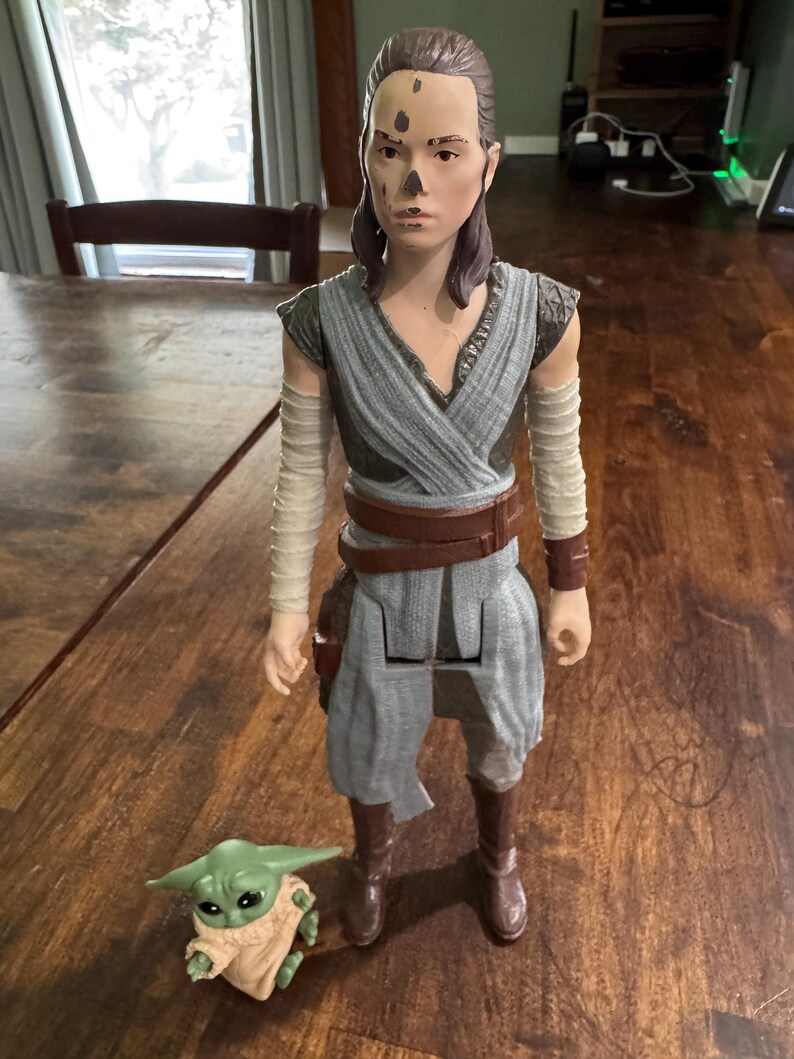 K&ouml;nnte beinhalten: Eine gro&szlig;e Actionfigur eines Charakters aus dem Star Wars-Franchise, die ein grau-blaues Outfit mit braunen Stiefeln und einem G&uuml;rtel tr&auml;gt. Eine kleine gr&uuml;ne Figur von Grogu befindet sich zu F&uuml;&szlig;en der Figur. Die Figuren stehen auf einer Holzoberfl&auml;che.