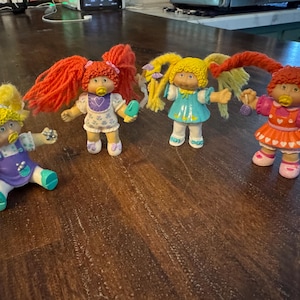 May include: Four Cabbage Patch Kids dolls with yarn hair and plastic bodies. One sits wearing a purple outfit, while three stand in colorful dresses. Each doll features distinct hair colors, including bright orange and yellow, and small accessories.