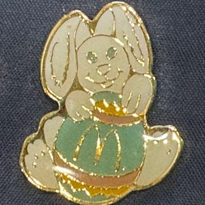 May include: A gold-colored pin featuring a white bunny holding a green and gold Easter egg. The bunny has a happy expression and is sitting on its hind legs.