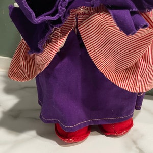 Puede incluir: Un vestido de mu&ntilde;eca a rayas rojas y moradas con zapatos rojos. El vestido tiene una falda morada y un corpi&ntilde;o a rayas rojas y blancas.