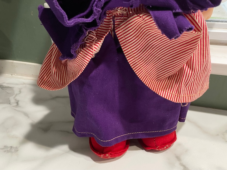 Puede incluir: Un vestido de mu&ntilde;eca a rayas rojas y moradas con zapatos rojos. El vestido tiene una falda morada y un corpi&ntilde;o a rayas rojas y blancas.