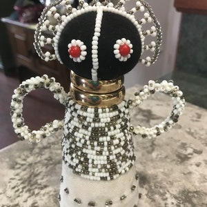 May include: A beaded doll with a black fabric head, white beads, and red accents. The doll has a white fabric skirt and gold rings around its neck.
