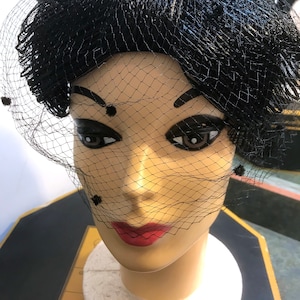 Puede incluir: Un sombrero negro con velo y red negra. El sombrero está hecho de un material suave y flexible y tiene una forma redondeada. El velo está unido al sombrero y cae sobre la cara. La red es fina y delicada.