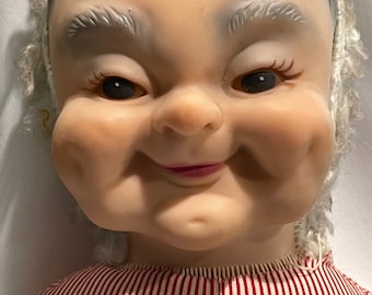 May include: A vintage doll with a mischievous grin, wearing a red and white striped dress. The doll has a realistic face with rosy cheeks and gray hair.