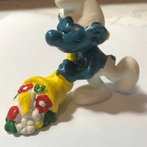 May include: A blue Smurf figurine holding a yellow cornucopia filled with red, white, and green flowers.