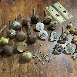 May include: A collection of antique door knobs and handles in various styles and finishes, including brass, glass, and porcelain. Some knobs have intricate designs, while others are simple and elegant. The collection also includes two brass door plates.