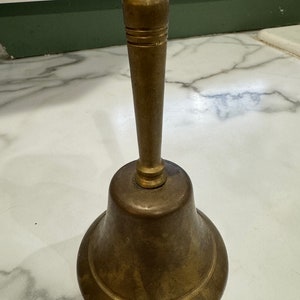 May include: A brass bell with a rounded, flared base and a tall, slender handle. The bell has a worn, aged finish.