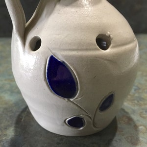 Puede incluir: Una jarra de cer&aacute;mica blanca con un dise&ntilde;o floral azul. La jarra tiene un asa y un pico. Hay agujeros en el lado de la jarra.