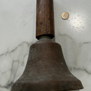 May include: A vintage copper bell with a wooden handle. The bell is tarnished and has a worn appearance. The handle is made of dark wood and has a slightly rough texture.