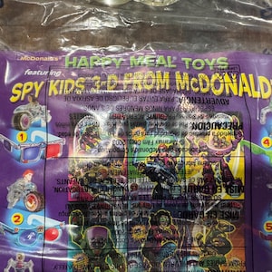 May include: A sealed Happy Meal toy bag featuring Spy Kids characters and accessories. The purple bag has the McDonald's logo and text. The bag contains small plastic toys, including figures and vehicles.