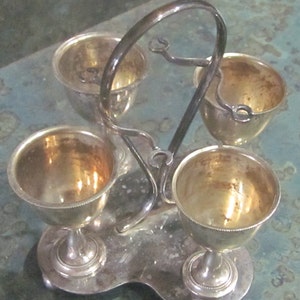 May include: Silver egg cup holder with three individual cups. The holder has a decorative handle and a base with a scalloped edge.