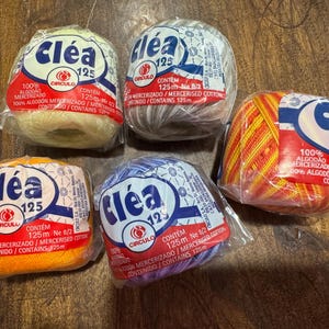May include: Five spools of Cléa 125 mercerized cotton yarn in various colors, including yellow, gray, orange, purple, and a mix of red and yellow. Each spool is wrapped in clear plastic and features a red and white label with the brand logo and product information.