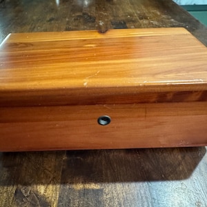 May include: A rectangular wooden box with a warm, reddish-brown finish. The box has a simple design with a small, round keyhole on the front. The wood grain is visible, adding to its natural aesthetic. The box is likely used for storage.