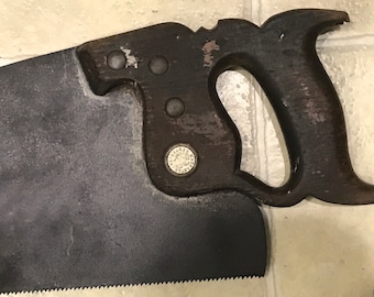 Könnte beinhalten: Eine antike Handsäge mit einem Holzgriff und einer Metallklinge. Der Griff hat eine Metallplatte mit den Worten "Disston" und "Made in USA" darauf.