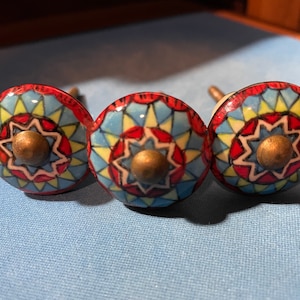 May include: Three ceramic cabinet knobs with a colorful geometric design. The knobs feature a starburst pattern in shades of red, blue, yellow, and white. Each knob has a brass center.