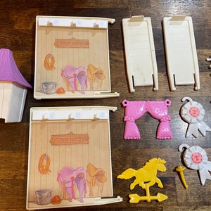 May include: A vintage My Little Pony playset with a purple stable, two tack rooms, a yellow horse, and various accessories. The tack rooms have a sign that reads "Tack Room".
