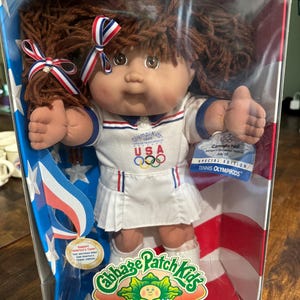 May include: A Cabbage Patch Kids doll, Carmella Nell, in a clear plastic box. The doll is wearing a white tennis outfit with red and blue accents and the Olympic rings. The box has a red, white, and blue theme with the words "OLYMPIKIDS" and "SPECIAL EDITION".