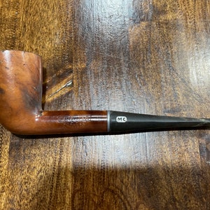 May include: A brown wooden pipe with a black stem and a silver band. The band has the letters "MC" engraved on it. The pipe is made by "Mastercraft" and is a "Popular" model.