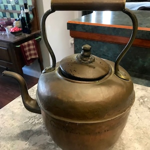 May include: A large, antique copper kettle with a wooden handle and a brass lid. The kettle has a rounded body and a spout on the side. The kettle is sitting on a light gray countertop.