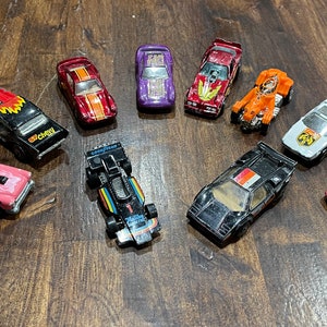 May include: A collection of vintage toy cars, including a pink car, a red car, a black car, a white car, a purple car, an orange car, a black and red car, a black and white car, a red and blue car, and a silver car.