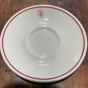 May include: White ceramic saucer with a red rim and a red logo in the center. The logo is a shield with the letter "I" inside.