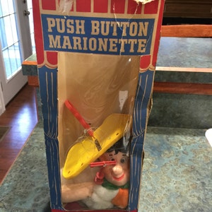 May include: A vintage cardboard box for a push button marionette toy. The box is red, white, and blue with the words "Push Button Marionette" printed on the front. The box features a clear plastic window showing a yellow hat, a red and white stick figure, and a smiling face with a red nose.