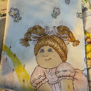 May include: Light blue Cabbage Patch Kids sheet featuring a cartoon doll with blonde pigtails tied with pink bows. The doll is wearing a pink dress. Scattered blue flowers and a yellow rainbow are also visible.