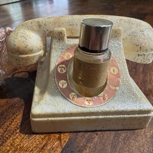 May include: A vintage-style telephone with a bottle of perfume resting on the dial. The phone is off-white with silver glitter and a pink tassel. The perfume bottle has a silver cap and a brown label.