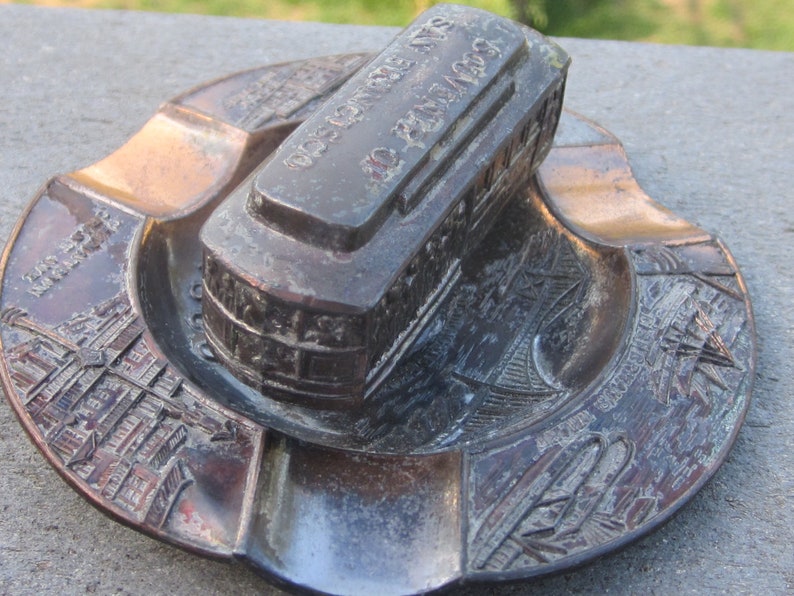 May include: A vintage metal ashtray featuring a detailed depiction of a San Francisco streetcar. The ashtray is decorated with the words "San Francisco, Calif. No. 2400, 1923" on the top of the streetcar. The ashtray is a great collectible for fans of San Francisco.