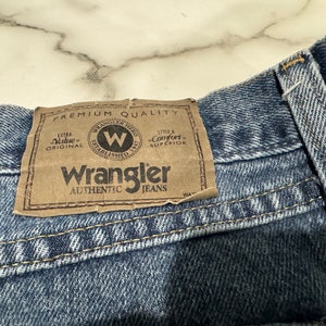 May include: A close-up of a blue denim Wrangler jean waistband with a brown leather patch that reads "Premium Quality", "Extra Value Original", "Wrangler Established 1947", "Style & Comfort Superior", and "Wrangler Authentic Jeans".