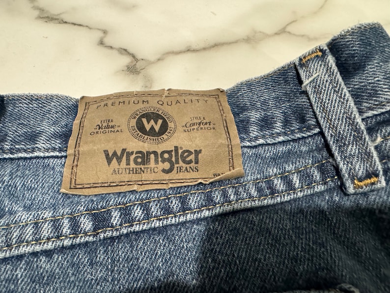 May include: A close-up of a blue denim Wrangler jean waistband with a brown leather patch that reads "Premium Quality", "Extra Value Original", "Wrangler Established 1947", "Style & Comfort Superior", and "Wrangler Authentic Jeans".