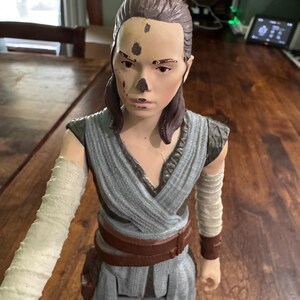 K&ouml;nnte beinhalten: Detaillierte Actionfigur einer weiblichen Figur, die ein graues und braunes Outfit mit einem braunen G&uuml;rtel tr&auml;gt. Die Figur hat einen hellen Teint, braune Haare und einen Arm mit wei&szlig;em Verband. Die Figur ist etwa 30 cm gro&szlig;.