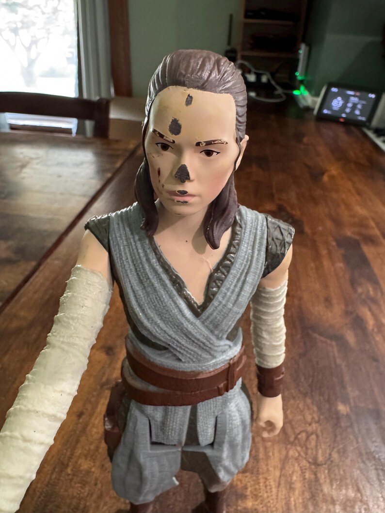 K&ouml;nnte beinhalten: Detaillierte Actionfigur einer weiblichen Figur, die ein graues und braunes Outfit mit einem braunen G&uuml;rtel tr&auml;gt. Die Figur hat einen hellen Teint, braune Haare und einen Arm mit wei&szlig;em Verband. Die Figur ist etwa 30 cm gro&szlig;.