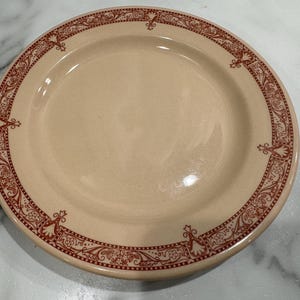 May include: A cream-colored ceramic plate with a red and white floral border design.