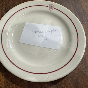 May include: Off-white ceramic plate with a red rim and a small logo. A handwritten note taped to the plate reads "Homer Laughter Small nic 7". The plate is 7 inches in diameter.