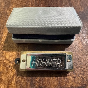 May include: A Hohner harmonica with a silver plate and the brand name in bold letters. The harmonica is gold-toned and rests on a wooden surface next to a gray box with a dark blue interior.
