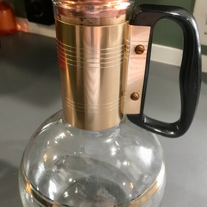 May include: A clear glass decanter with a gold band around the bottom and a copper colored lid. The decanter has a black plastic handle.