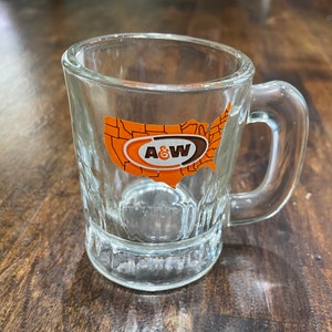 May include: A clear glass mug with a handle. The mug has a raised design and features a logo of A&W with a stylized map of the United States.