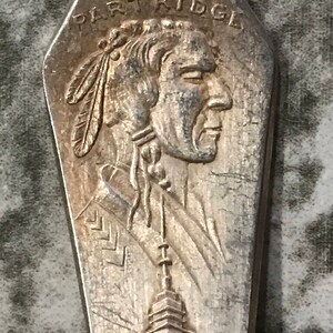 May include: Silver-toned metal spoon with a profile of a Native American man with a feather headdress and the text "BLACK PARTRIDGE" above. The spoon also features a stylized image of a building.