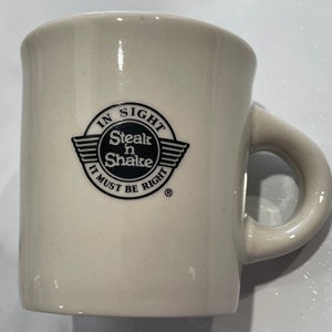 May include: Off-white ceramic mug with a rounded handle. The mug features the Steak 'n Shake logo in black, with the text "IN SIGHT Steak n Shake IT MUST BE RIGHT". The mug is likely for beverages.