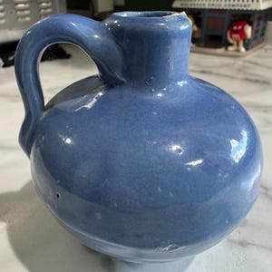 May include: A blue ceramic jug with a rounded body and a handle. The jug has a smooth, glossy finish and a small opening at the top. The jug is a light blue color and appears to be vintage.