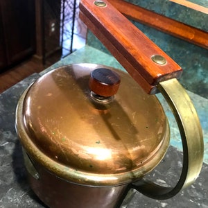 May include: A copper pot with a wooden handle and a brass lid. The pot has a brass band around the top and a brass handle on the side.