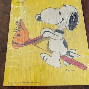 May include: A yellow wooden puzzle featuring a cartoon image of Snoopy riding a red and orange rocking horse. The puzzle is made by Playskool and includes the text "Snoopy" Copr. 1958 United Feature Syndicate, Inc. and "SCHULZ".