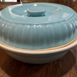 May include: A light blue ceramic casserole dish with a matching lid. The dish has a ribbed design around the sides and a small handle on the lid. The dish sits on a white rimmed base.