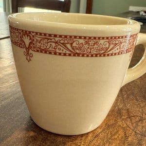 May include: A white ceramic coffee mug with a red and white floral design around the top rim. The mug has a rounded shape and a handle.