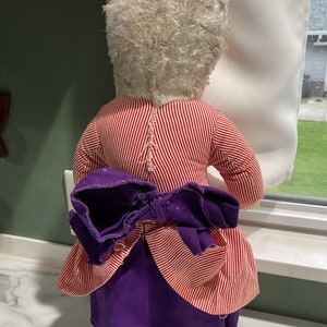 Puede incluir: Una mu&ntilde;eca vintage con pelo blanco, vestida con un vestido a rayas rojas y blancas con una falda morada y un lazo morado. La mu&ntilde;eca est&aacute; de pie sobre una superficie blanca.