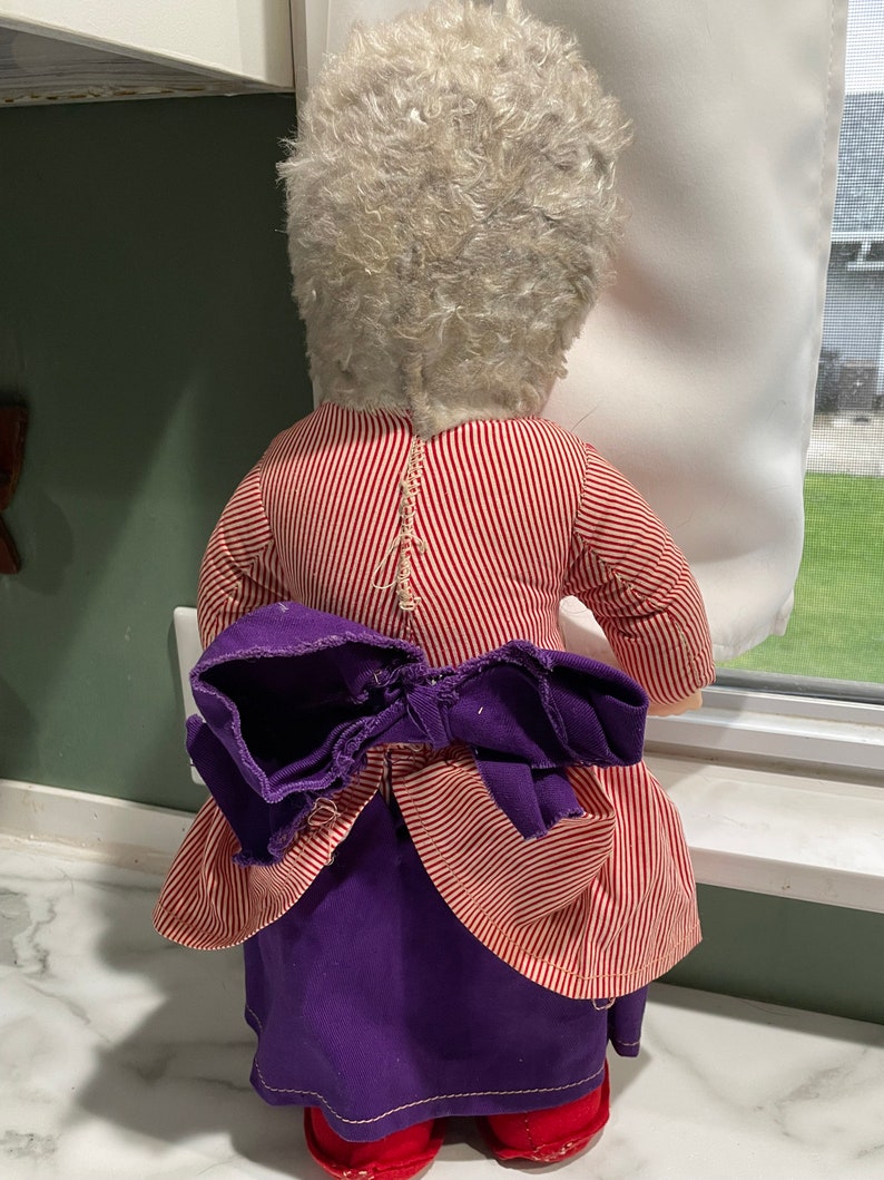 Puede incluir: Una mu&ntilde;eca vintage con pelo blanco, vestida con un vestido a rayas rojas y blancas con una falda morada y un lazo morado. La mu&ntilde;eca est&aacute; de pie sobre una superficie blanca.