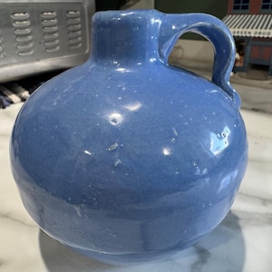 May include: A round, light blue ceramic jug with a handle. The jug has a speckled finish and a rounded body. The handle is arched and attached to the top of the jug. The jug is sitting on a marble surface.