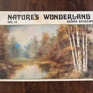 May include: A vintage book cover titled "Nature's Wonderland Vol. 13" by Susan Scheewe. The cover features a landscape painting of a serene lake surrounded by trees with fall foliage. The color palette includes browns, greens, and blues.