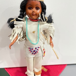 May include: A vintage Native American doll wearing a white leather outfit with fringe, a turquoise beaded necklace, and a brown headband with red flowers. The doll has black hair in braids and brown eyes.